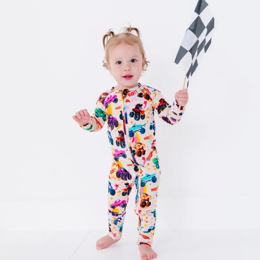 A baby standing in a cream zip-up pajama with a colorful monster truck print, holding a checkered racing flag.